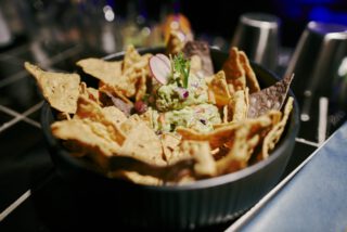 Teller mit Guacamole, Tortilla‑Chips in dunklem Mörser, samt Getränkeglas daneben.