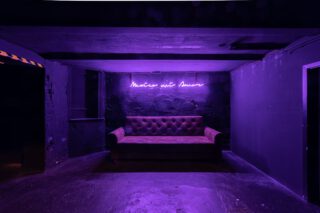 Lila beleuchtete Sitzecke mit abgenutztem Samtsofa vor einer schwarzen Wand und Neon-Schriftzug ‚México mit Amor‘ in Handschrift-Stil.