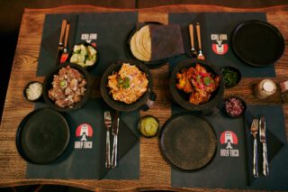 Gedeckter Holztisch im Holy Taco mit drei Molcajete-Schalen gefüllt mit verschiedenen Fleischsorten, dazu Limetten, Zwiebeln, Guacamole, frische Kräuter, Tortillas und Besteck auf schwarzen Platzsets mit Restaurantlogo.