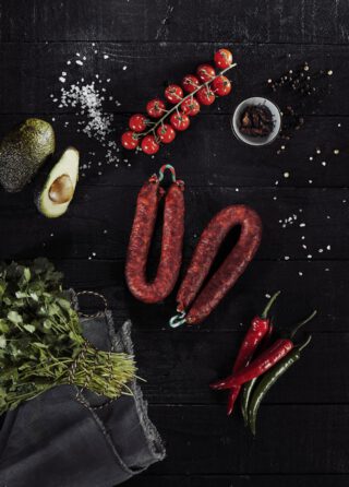Flach gelegte Zutaten auf schwarzem Holz: Zwei getrocknete Chorizo-Würste, rote und grüne Chilischoten, ein Bund frischer Koriander, halbierte Avocado, Cocktailtomaten an der Rispe, grobes Meersalz, Pfefferkörner und getrocknete Chilis in einer kleinen Schale.