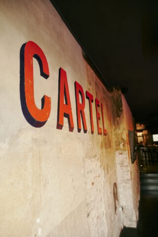 Verwitterte Backsteinwand im Restaurant mit großem, retrostilisiertem Schriftzug ‚CARTEL‘ in orange-roten 3D-Buchstaben, leicht perspektivisch fotografiert im schummrigen Eingangsbereich.