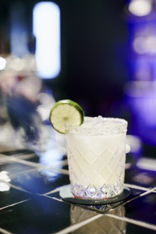 Cocktailglas mit Salzrand und Limettenscheibe auf schwarzer Fliesenbar im Holy Taco, gefüllt mit einem klaren Getränk vor unscharfem, stimmungsvoll beleuchtetem Hintergrund.