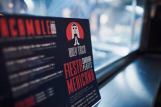 Menükarte des Holy Taco mit rotem Logo und der Aufschrift ‚Fiesta Mexicana – Sharing Plates‘ vor einem Fenster mit Tageslichtreflexion.