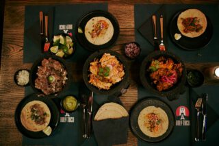 Von oben fotografierter Holztisch im Holy Taco, gedeckt mit verschiedenen Tacos, Molcajete-Schalen mit Fleischfüllungen, Beilagen wie Limetten, Zwiebeln und Guacamole sowie Besteck und dunklen Tellern.
