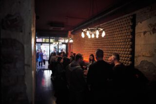 Abendliche Szene im Holy Taco: Menschen stehen und unterhalten sich an der Bar vor einer Wand aus goldfarbenen Totenköpfen, beleuchtet von industriellen Hängelampen; im Hintergrund Blick auf die geöffnete Eingangstür mit weiteren Gästen draußen.