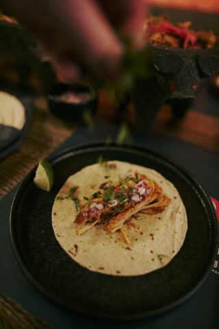 Frisch belegter Taco auf einem dunklen Teller, gefüllt mit Fleisch, roten Zwiebeln, Kräutern und Koriander; darüber streut eine Hand frische Zutaten, daneben liegt ein Stück Limette.