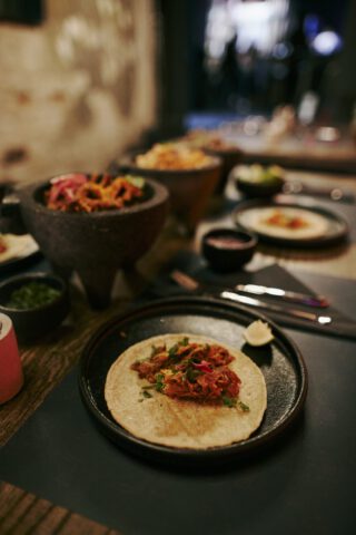 Taco auf dunklem Teller, gefüllt mit mariniertem Fleisch, Kräutern und Zwiebeln, daneben ein Stück Limette; im Hintergrund stehen weitere Molcajete-Schalen mit Beilagen auf einem gedeckten Holztisch im Restaurant.