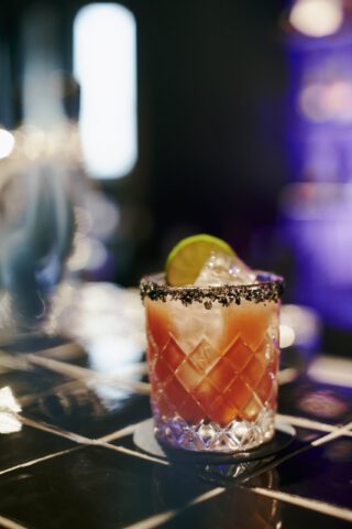 Nahaufnahme eines Cocktails in einem geschliffenen Glas mit schwarzem Salzrand und Limettenscheibe, serviert auf schwarzem Fliesentresen in stimmungsvoll beleuchteter Barumgebung.