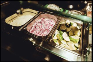 Edelstahl-Salatbar mit drei Behältern, gefüllt mit gewürfelten weißen Zwiebeln, roten Zwiebeln und frischen Limettenspalten.
