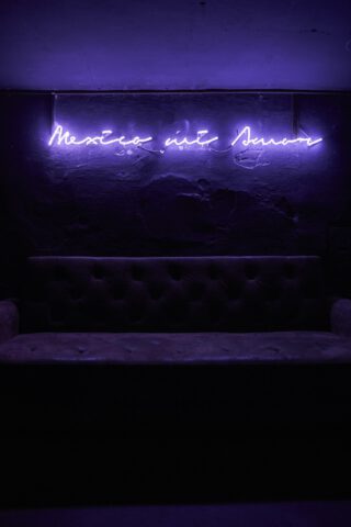 Dunkles Interieur mit violett leuchtendem Neonschriftzug ‚Mexico mit Amor‘ über einem abgenutzten, gepolsterten Sofa vor rauer Wand.