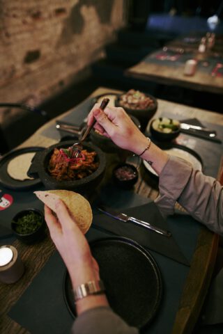 Gast sitzt an einem gedeckten Tisch im Restaurant und bereitet mit einer Gabel und einer Tortilla ein Taco-Gericht mit mariniertem Fleisch, eingelegten Zwiebeln und Beilagen in kleinen Schälchen zu.