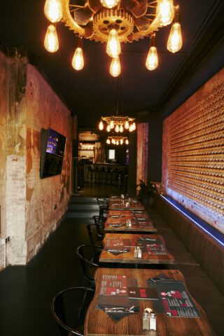 Innenraum des Restaurants Holy Taco mit rustikaler Wand, goldener Totenkopf-Installation, gedeckten Holztischen und warmem Licht von industriellen Deckenlampen – Blickrichtung zur Bar im Hintergrund.