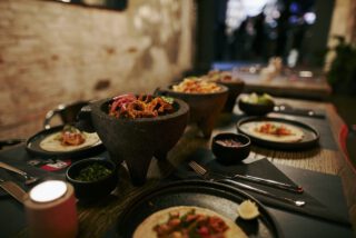 Rustikaler Holztisch im Restaurant, gedeckt mit Tacos, Limetten, Saucen und traditionellen Molcajete-Schalen mit verschiedenen Füllungen, in stimmungsvoll beleuchtetem Ambiente.