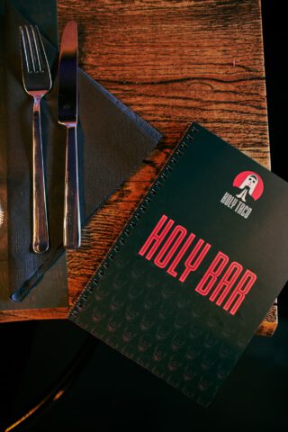 Nahaufnahme eines gedeckten Holztischs im Holy Taco mit Besteck auf schwarzer Serviette und einer Bar-Karte mit Totenkopf-Muster und der Aufschrift ‚HOLY BAR‘ in roter Schrift.
