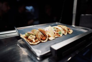 Sechs verschiedene Tacos auf einem Tablett in der Restaurantküche, abwechslungsreich gefüllt mit Gemüse, Fleisch, Salsa, Guacamole und Zwiebeln – stilvoll angerichtet unter hellem Licht.