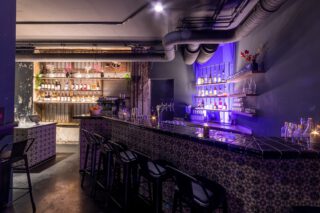 Stimmungsvoll beleuchtete Bar im Holy Taco mit Fliesenverkleidung, hohen Barhockern, modernen Spirituosenregalen und violett-blauer Ambientebeleuchtung, ergänzt durch sichtbare Lüftungsrohre und dekorative Details.