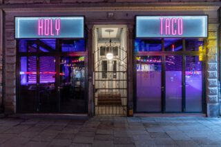 Abendliche Außenansicht des Restaurants 'Holy Taco' mit zwei leuchtenden Schildern über den Eingängen: links in pink 'HOLY', rechts 'TACO'. Zwischen den Eingängen befindet sich eine beleuchtete Altbau-Haustür mit Gitter. Im Inneren des Restaurants sind violette Neonlichter und bunte Dekorationen zu sehen.