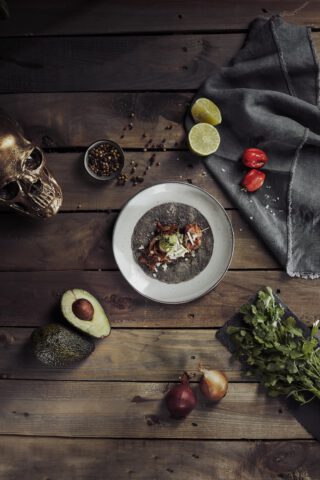 Ein einzelner Taco mit dunkler Maistortilla auf einem weißen Teller, serviert auf einem rustikalen Holztisch. Umgeben von halbierter Avocado, roten und gelben Zwiebeln, frischem Koriander, einer grauen Serviette, Limettenhälften, kleinen roten Chilis, gemischten Pfefferkörnern und einem goldfarbenen Totenschädel als Dekoration.