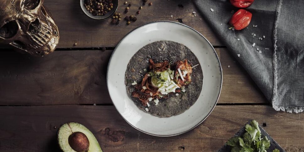 Ein einzelner Taco mit dunkler Maistortilla auf einem weißen Teller, serviert auf einem rustikalen Holztisch. Umgeben von halbierter Avocado, roten und gelben Zwiebeln, frischem Koriander, einer grauen Serviette, Limettenhälften, kleinen roten Chilis, gemischten Pfefferkörnern und einem goldfarbenen Totenschädel als Dekoration.