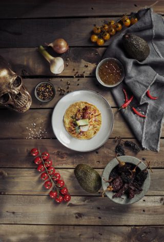 Teller mit einem belegten Taco auf rustikalem Holztisch, umgeben von Knoblauch, Zwiebel, Avocados, roten und gelben Kirschtomaten, Chilischoten, getrockneten Chilis in einer Steinmörserschale, Schale mit Salsa, Pfefferkörnern, Salz und einem goldfarbenen Totenschädel als Dekoration.