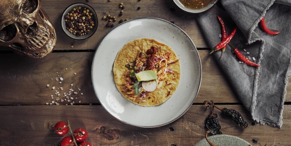 Teller mit einem belegten Taco auf rustikalem Holztisch, umgeben von Knoblauch, Zwiebel, Avocados, roten und gelben Kirschtomaten, Chilischoten, getrockneten Chilis in einer Steinmörserschale, Schale mit Salsa, Pfefferkörnern, Salz und einem goldfarbenen Totenschädel als Dekoration.