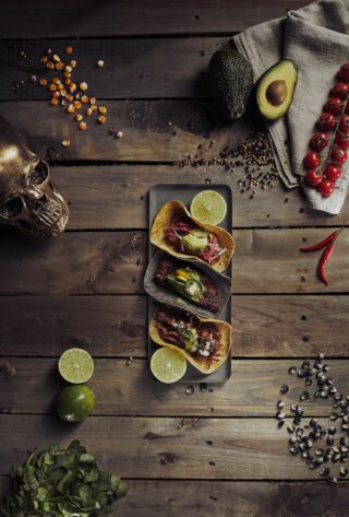 Drei belegte Tacos auf einem dunklen Teller, serviert auf einem rustikalen Holztisch, garniert mit Limettenhälften. Umgeben von Avocados, Cherrytomaten am Zweig, Knoblauch, roten Chilischoten, schwarzen und gelben Pfefferkörnern, Maiskörnern, schwarzen Maiskörnern, frischem Koriander, einem beigen Küchentuch und einem dekorativen goldfarbenen Totenschädel.