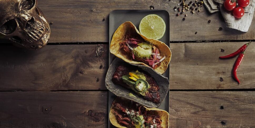 Drei belegte Tacos auf einem dunklen Teller, serviert auf einem rustikalen Holztisch, garniert mit Limettenhälften. Umgeben von Avocados, Cherrytomaten am Zweig, Knoblauch, roten Chilischoten, schwarzen und gelben Pfefferkörnern, Maiskörnern, schwarzen Maiskörnern, frischem Koriander, einem beigen Küchentuch und einem dekorativen goldfarbenen Totenschädel.
