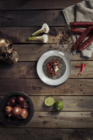 Teller mit einem Taco auf einer dunklen Tortilla, garniert mit Zwiebeln, Fleisch und Limette, angerichtet auf einem rustikalen Holztisch mit Knoblauch, Limetten, roten Zwiebeln, getrockneter Wurst, Chilischoten, Gewürzen und einem dekorativen Totenkopf.