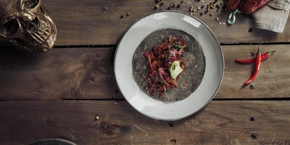 Teller mit einem Taco auf einer dunklen Tortilla, garniert mit Zwiebeln, Fleisch und Limette, angerichtet auf einem rustikalen Holztisch mit Knoblauch, Limetten, roten Zwiebeln, getrockneter Wurst, Chilischoten, Gewürzen und einem dekorativen Totenkopf.