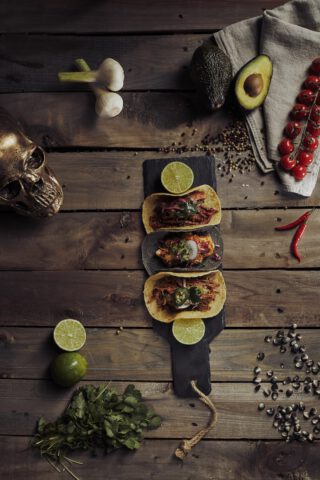 Drei Tacos auf einem dunklen Servierbrett mit Limettenscheiben, angerichtet auf einem rustikalen Holztisch, umgeben von Knoblauch, Avocado, Kirschtomaten, Chilischoten, Pfefferkörnern, Koriander, einer Limette, Maiskörnern und einem dekorativen Totenkopf.