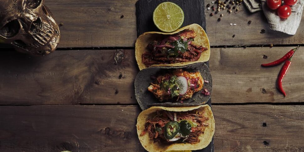 Drei Tacos auf einem dunklen Servierbrett mit Limettenscheiben, angerichtet auf einem rustikalen Holztisch, umgeben von Knoblauch, Avocado, Kirschtomaten, Chilischoten, Pfefferkörnern, Koriander, einer Limette, Maiskörnern und einem dekorativen Totenkopf.