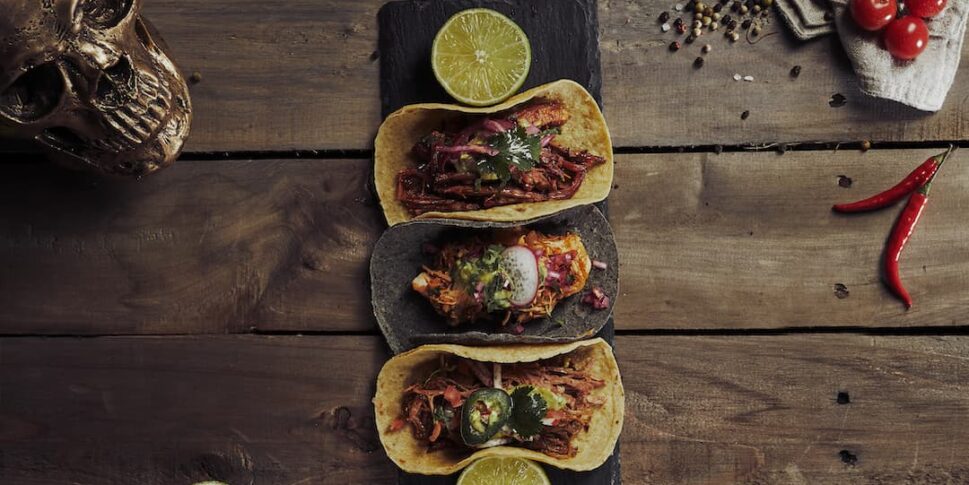Drei Tacos auf einem schwarzen Servierbrett mit Limettenscheiben, angerichtet auf rustikalem Holztisch. Die Tacos sind mit Fleisch, roten Zwiebeln, Koriander, Jalapeños und Radieschenscheiben gefüllt. Umgeben von einer halbierten Avocado, Kirschtomaten am Zweig, ganzen Limetten, roten Chilischoten, Pfefferkörnern, Maiskörnern, Kräutern und einem dekorativen Metallschädel.