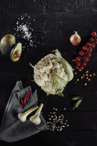 Ein ganzer Blumenkohl liegt auf einem dunklen Holzuntergrund, umgeben von einer halbierten Avocado, einer Zwiebel, einer Rispe roter Kirschtomaten, roten Chilischoten, zwei Knoblauchzehen auf einem grauen Tuch, Maiskörnern und einigen grünen Jalapeños.