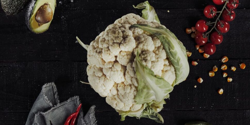 Ein ganzer Blumenkohl liegt auf einem dunklen Holzuntergrund, umgeben von einer halbierten Avocado, einer Zwiebel, einer Rispe roter Kirschtomaten, roten Chilischoten, zwei Knoblauchzehen auf einem grauen Tuch, Maiskörnern und einigen grünen Jalapeños.