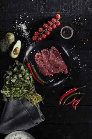 Drei rohe Rindersteaks mit grobem Meersalz auf einem schwarzen Teller, platziert auf einem dunklen Holzuntergrund. Umgeben von einer halbierten Avocado, einem Bund frischem Koriander, roten und grünen Chilischoten, einer Rispe Kirschtomaten, Pfefferkörnern und einer kleinen Schale mit getrockneten Chilis.