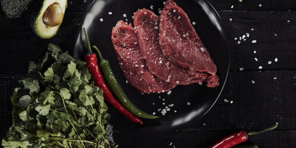 Drei rohe Rindersteaks mit grobem Meersalz auf einem schwarzen Teller, platziert auf einem dunklen Holzuntergrund. Umgeben von einer halbierten Avocado, einem Bund frischem Koriander, roten und grünen Chilischoten, einer Rispe Kirschtomaten, Pfefferkörnern und einer kleinen Schale mit getrockneten Chilis.