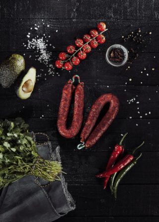 Zwei luftgetrocknete, rote Würste liegen auf einem dunklen Untergrund, umgeben von Kirschtomaten an der Rispe, halbierter Avocado, frischem Koriander, roten und grünen Chilischoten, grobem Salz und einer kleinen Schale mit getrockneten Chilistücken.