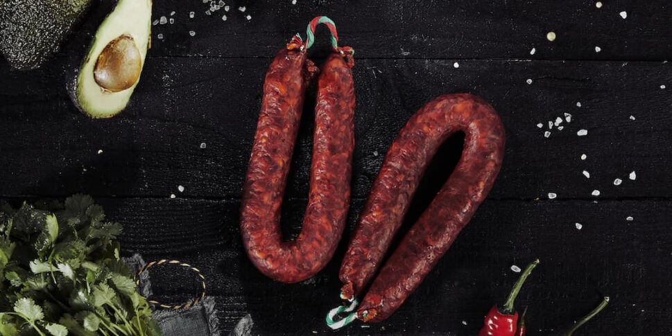 Zwei luftgetrocknete, rote Würste liegen auf einem dunklen Untergrund, umgeben von Kirschtomaten an der Rispe, halbierter Avocado, frischem Koriander, roten und grünen Chilischoten, grobem Salz und einer kleinen Schale mit getrockneten Chilistücken.