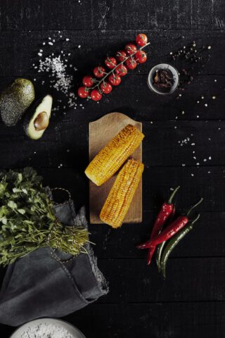 Zwei gegrillte Maiskolben auf einem Holzbrett, umgeben von frischen Zutaten wie Avocado, Kirschtomaten, Koriander, Chilis, Salz und Gewürzen auf dunklem Holzhintergrund.